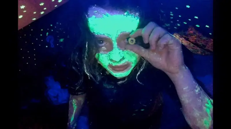 A closeup image of Casey the protaganist of We’re All Going to the World’s Fair with her face covered in something black light reactive, holding something up to her eye and just generally looking like a creep.