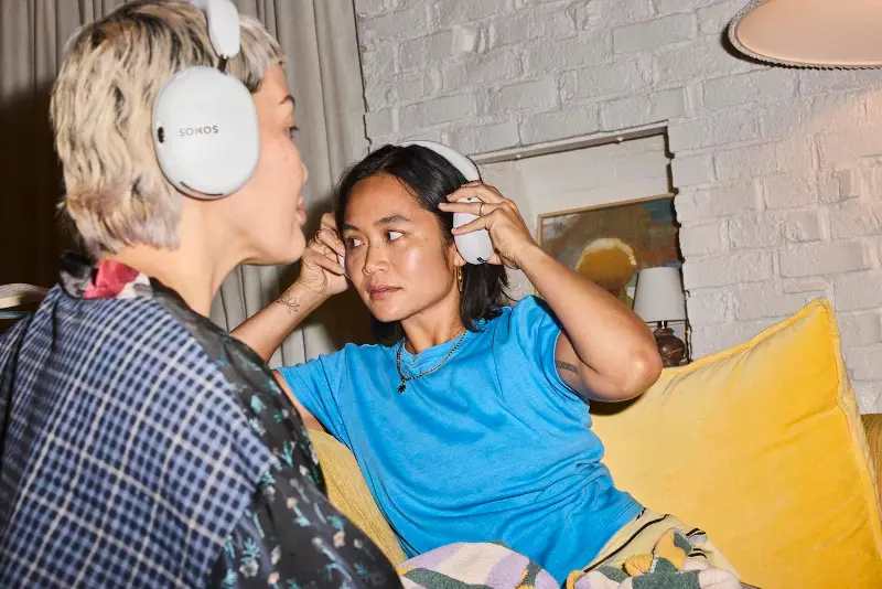 Two people sitting on a couch wearing the Sonos Ace Ultra headphones.