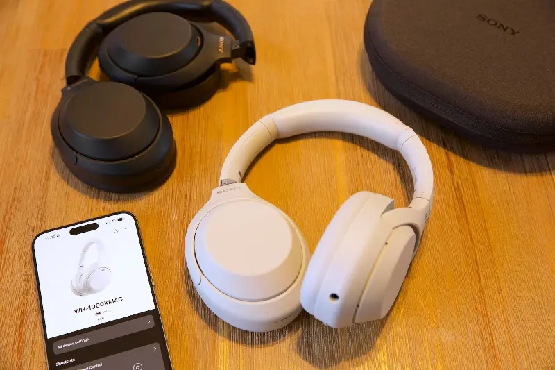 The white Sony WH-1000XM4C headphones next to black WH-1000XM4 headphones, an iPhone, and their black carrying case on a wooden table.