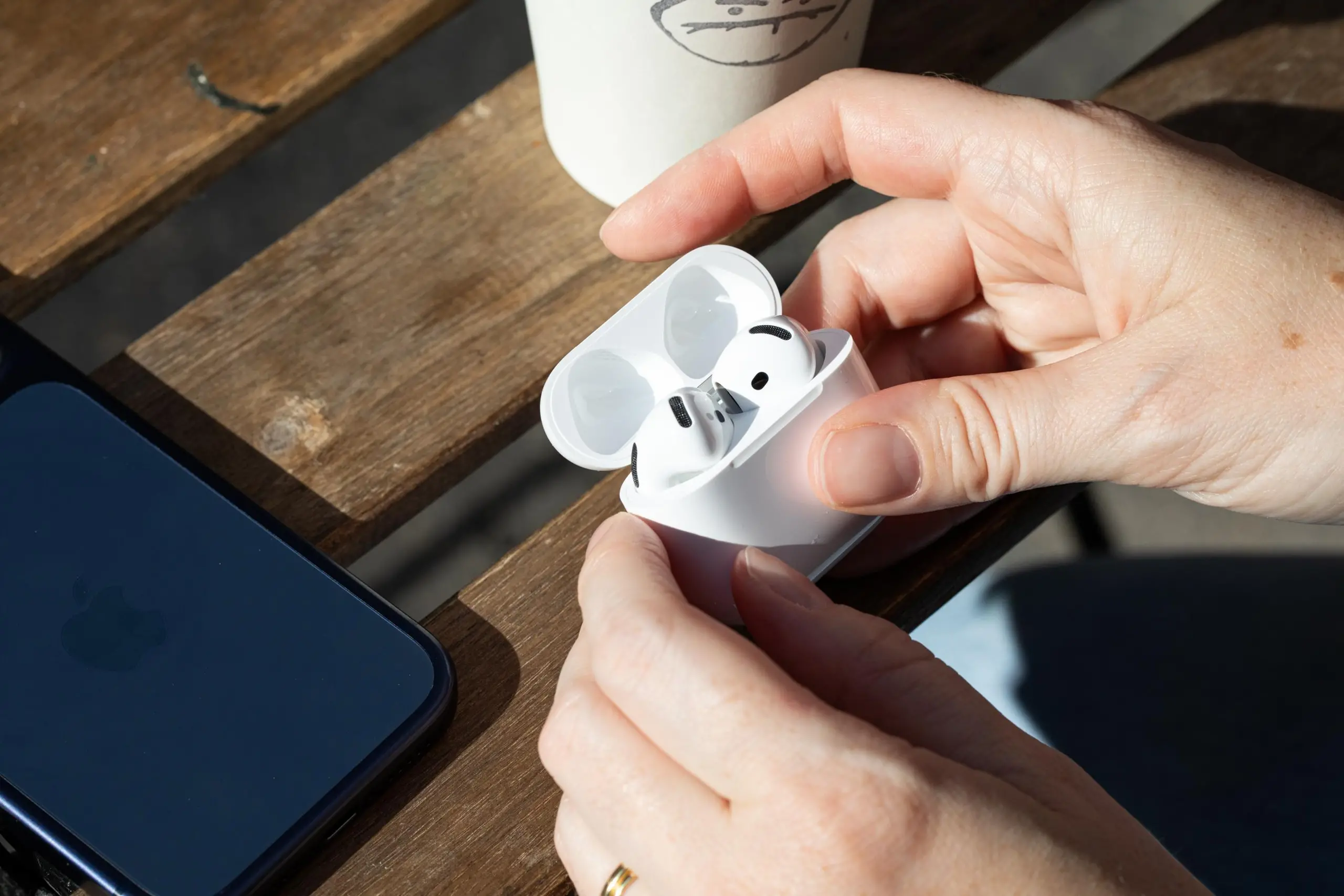 Hands holding the Apple AirPods 5 earbuds above a wooden-slated table with an iPhone and coffee cup on it.
