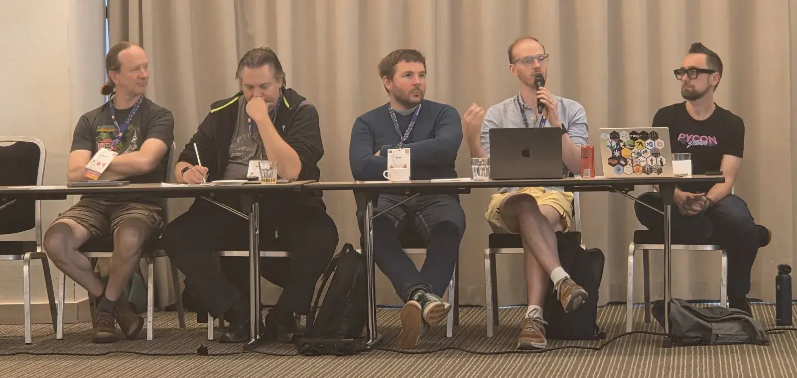David Hewitt speaking into a microphone, seated in a row of attendees