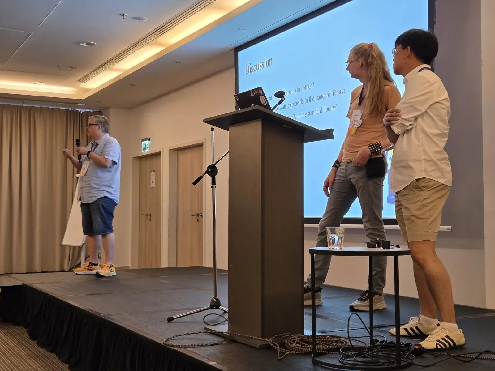 Tobias Wrigstad answering questions with a microphone on stage, with Fridtjof Stoldt and Donghee Na beside him