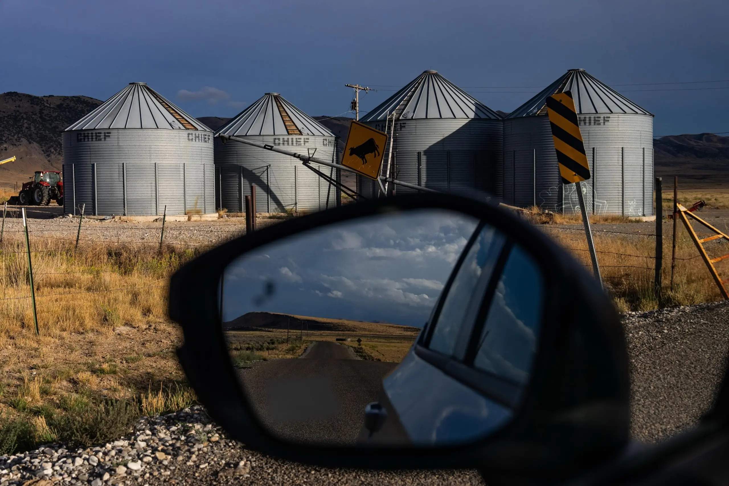 View of grain silos from the perspective of a driver looking at a rearview mirror, showing the road behind them in the reflection.