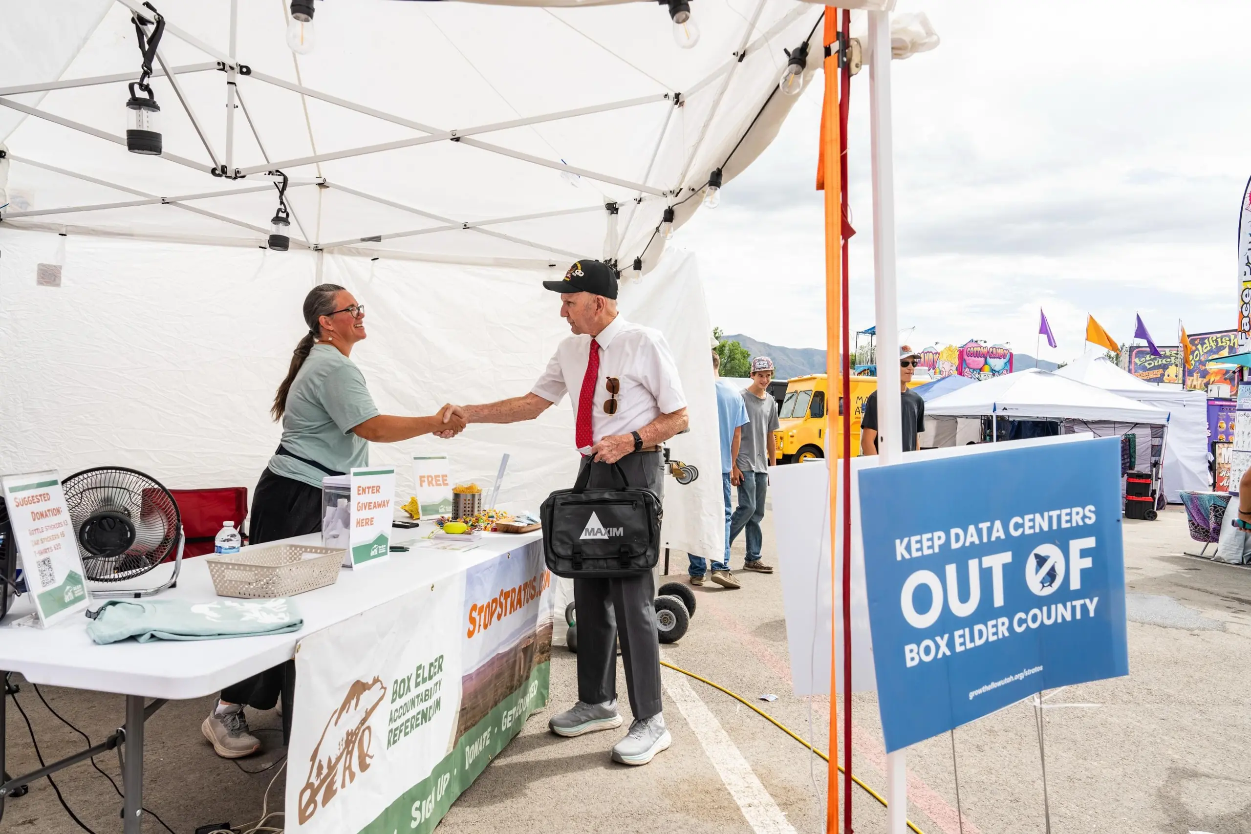 A man and a woman shaking hands beneath a pop-up tent with a sign in the foreground that reads "keep data centers out of Box Elder County."