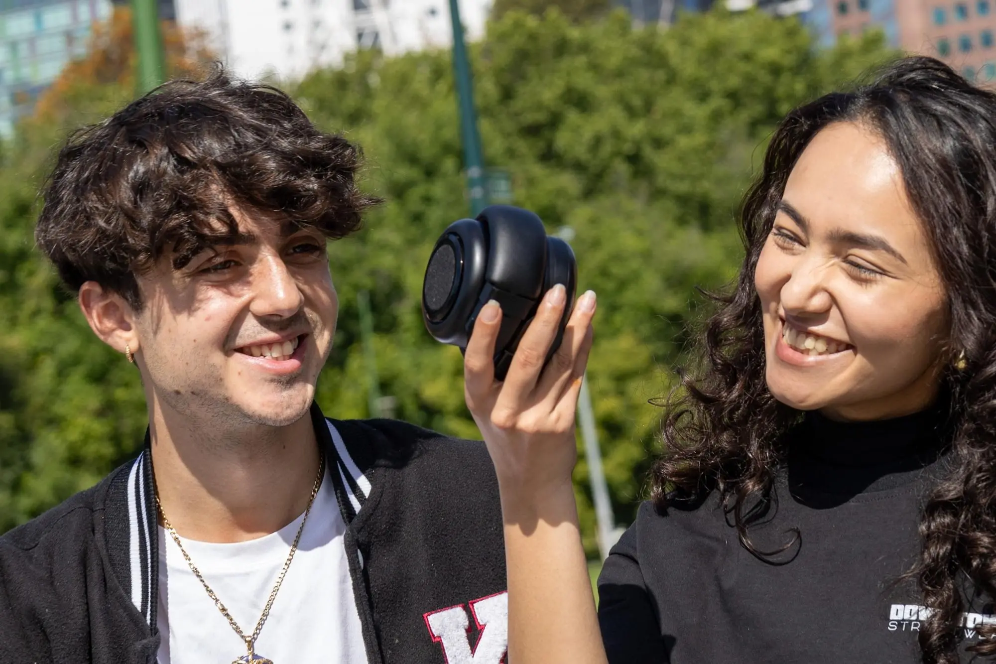 Two people sitting on an outdoor bench while one of them holds the TDM Neo headphones rolled up into Bluetooth speaker mode.