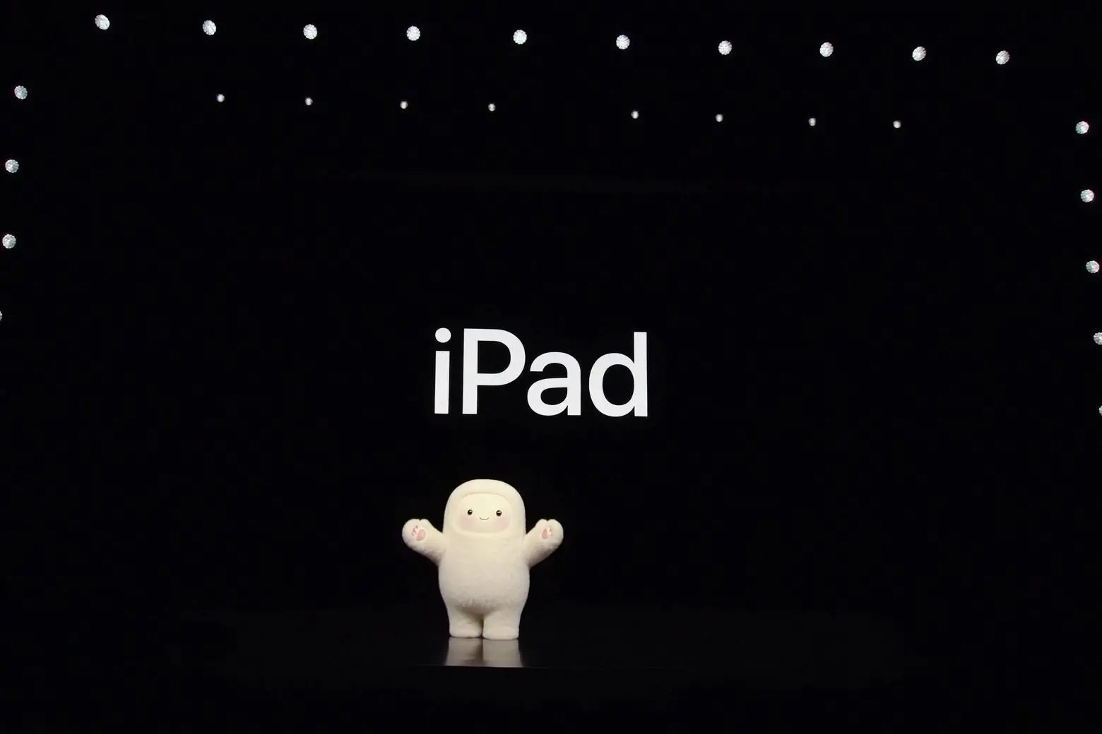 The Meta Muse mascot standing on a stage surrounded by lights with the word iPad on screen behind it.