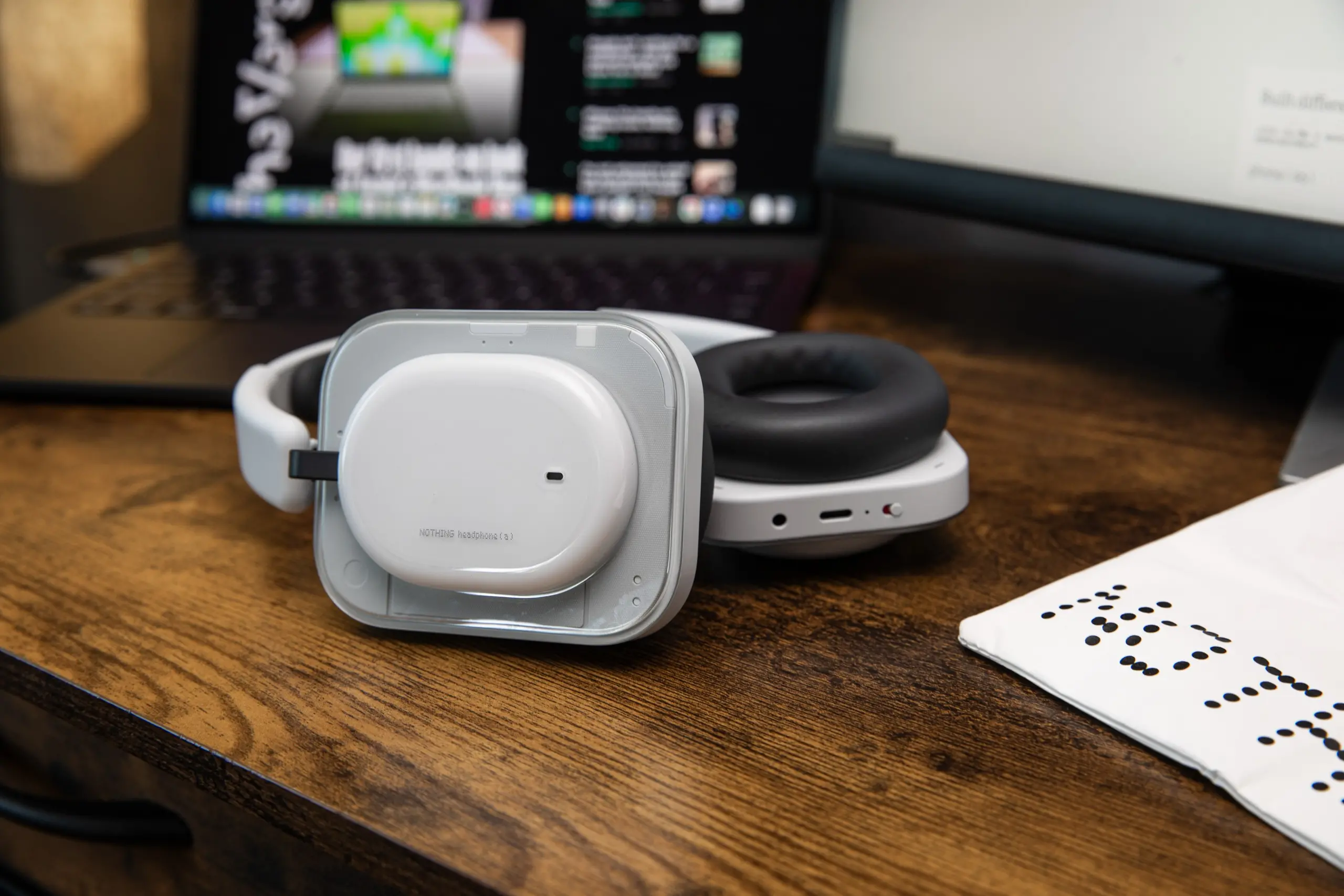 The Nothing Headphone A in white on a wooden desk with the carry bag off to the side and an open laptop behind.