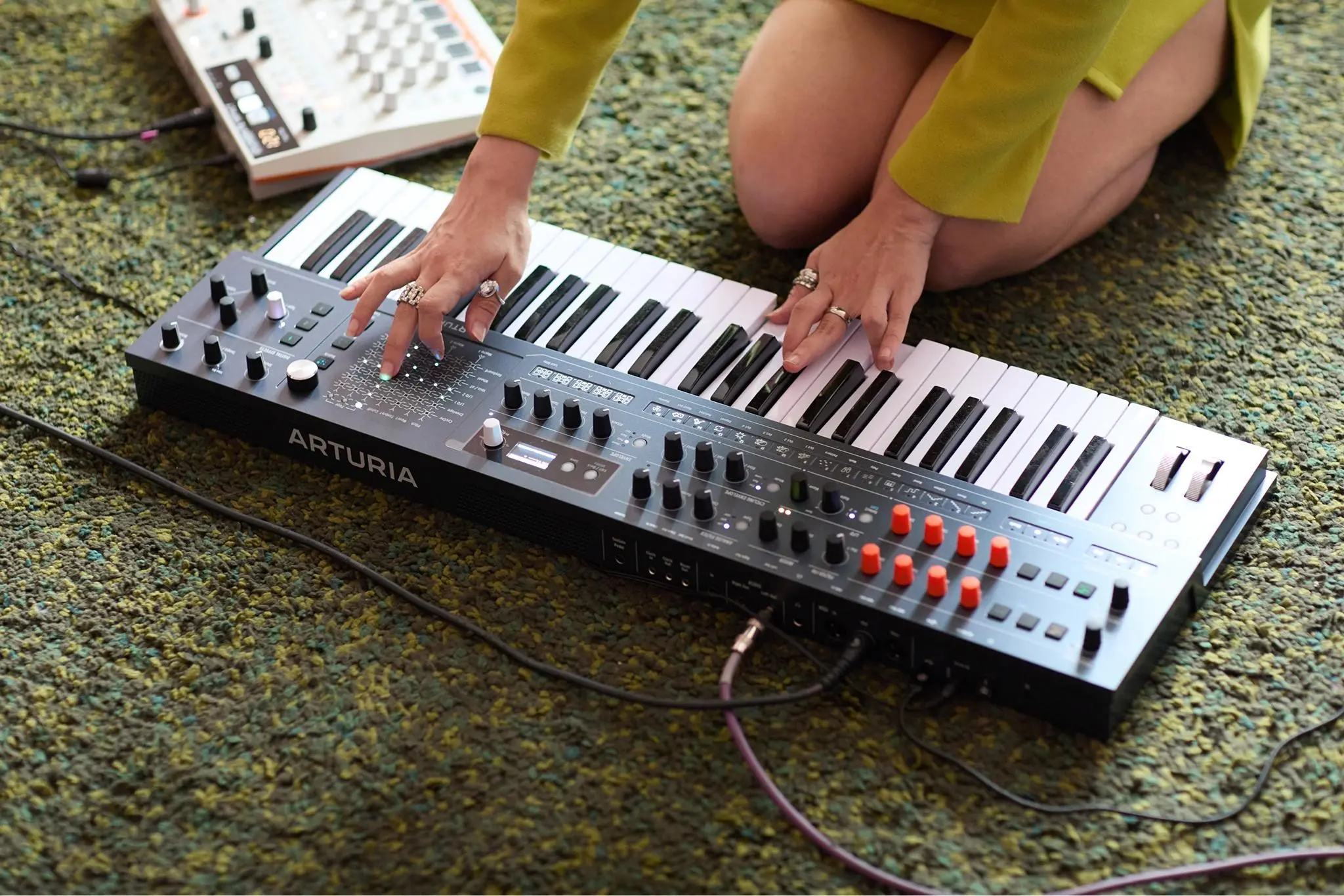 A woman kneeling on a green rug playing the Arturia MiniFreak 49 and using its XYZ touchpad.