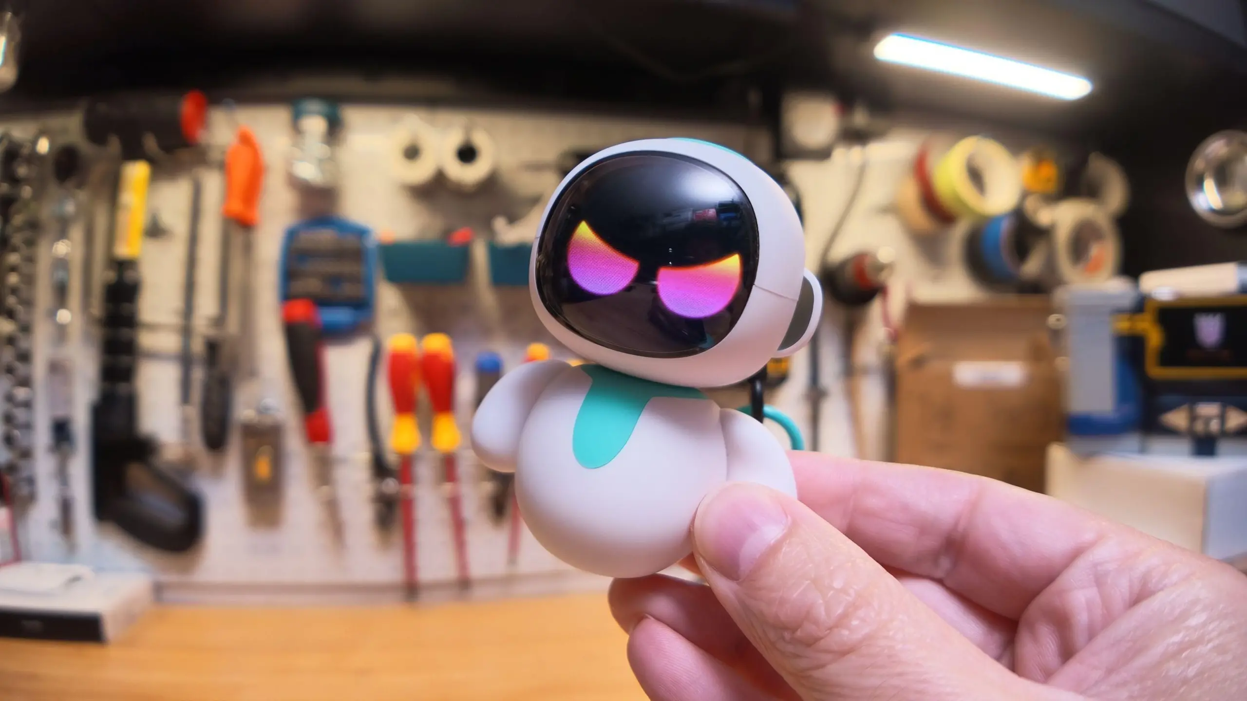 A cute little robot toy with scary rainbow eyes, held with my thumb and forefinger, with my garage workbench in the background.