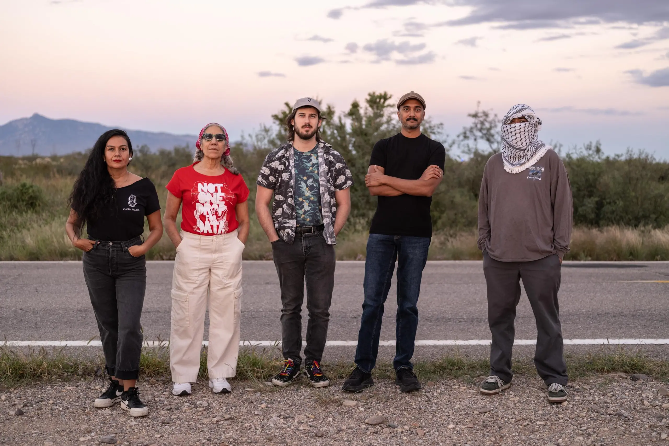 Five people standing side by side next to a desert road close to sunset.